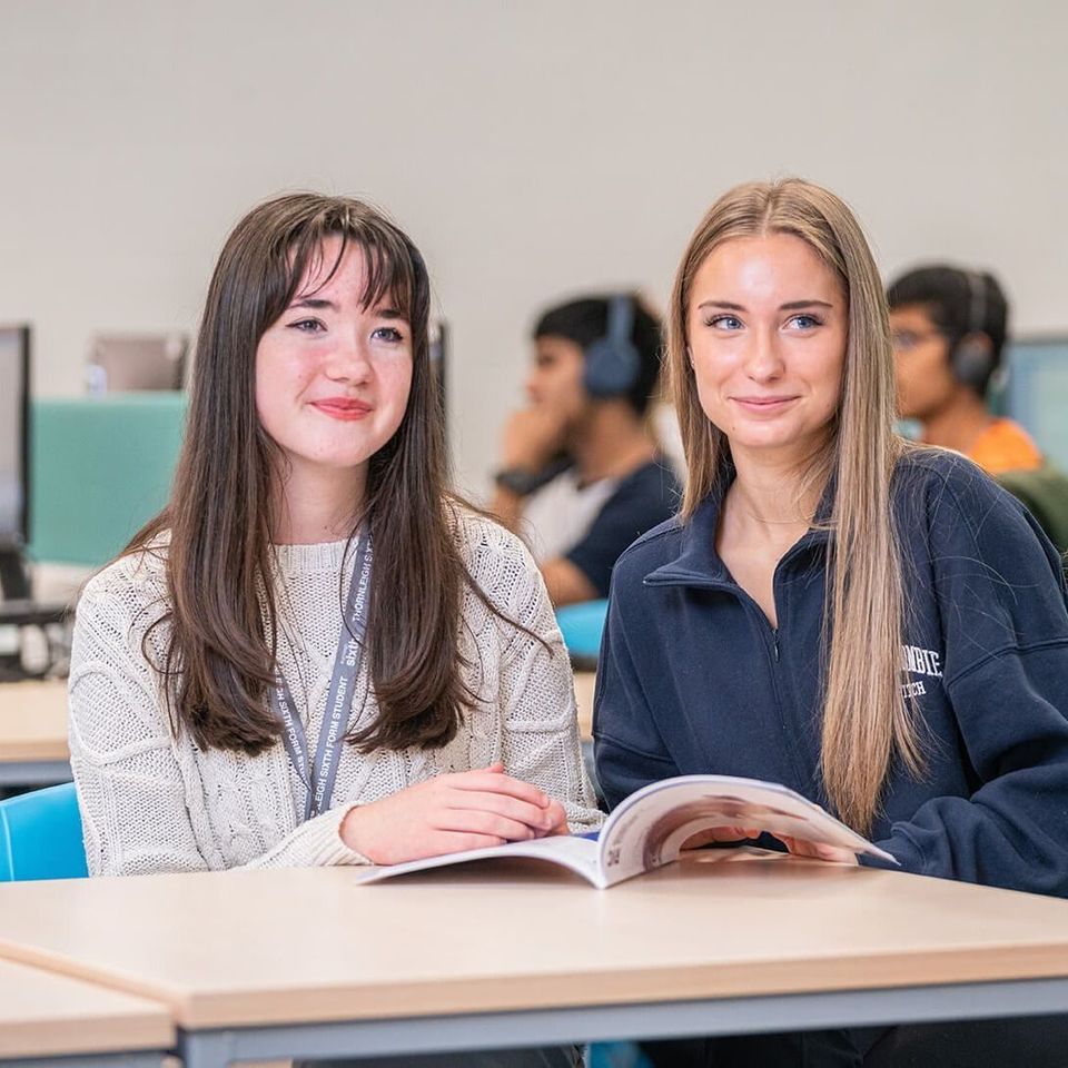 Two people are sitting at a table in a classroom, smiling and looking at a book. Two people are sitting at a table in a classroom, smiling and looking at a book.